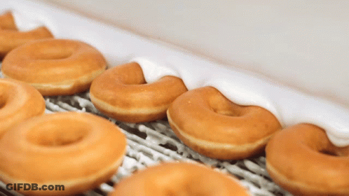 A Row Of Glazed Donuts With White Frosting On A Conveyor Belt GIF
