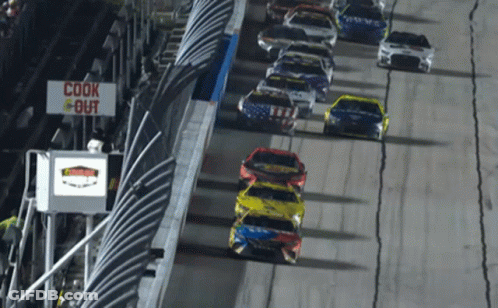 A Row Of Race Cars Are Driving Down A Track With A Sign That Says Cook Out GIF