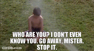 A Shirtless Boy Is Sitting In The Grass With A Sprinkler Spraying Water On Him . GIF