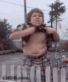 A Shirtless Boy Is Standing In Front Of A Fence And Making A Funny Face . GIF