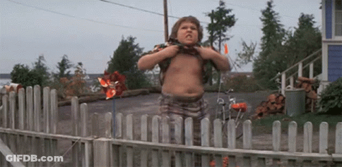 A Shirtless Boy Is Standing In Front Of A Wooden Fence With A Pinwheel In The Background . GIF
