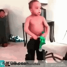 A Shirtless Little Boy Is Standing In A Living Room Holding A Green Toy . GIF
