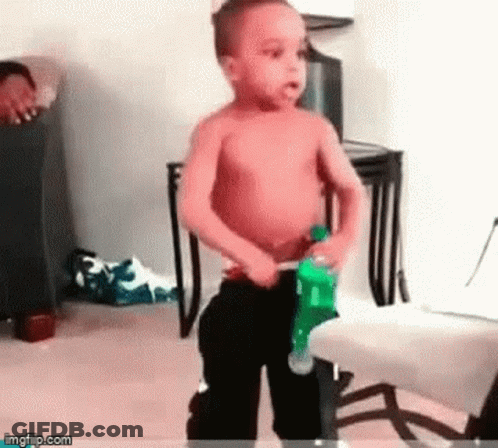 A Shirtless Little Boy Is Standing In A Room Holding A Bottle Of Soda . GIF