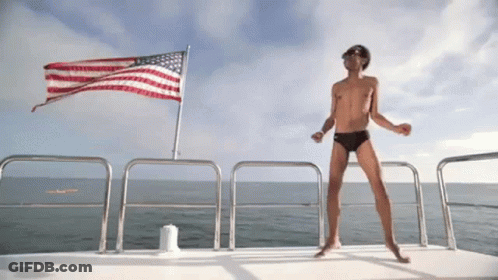 A Shirtless Man Dancing On A Boat In Front Of An American Flag GIF