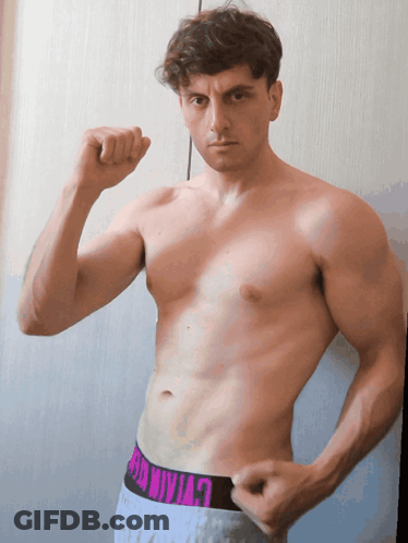 A Shirtless Man Flexes His Muscles In Front Of A Wall GIF