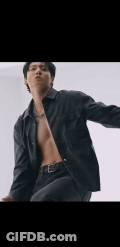 A Shirtless Man In A Black Jacket Is Standing In Front Of A White Wall With His Arms Outstretched . GIF
