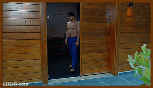 A Shirtless Man In Blue Pants Stands In The Doorway Of A Wooden House GIF