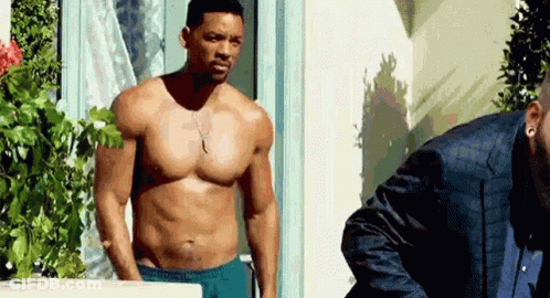 A Shirtless Man In Blue Shorts Is Standing Next To Another Man In A Suit . GIF