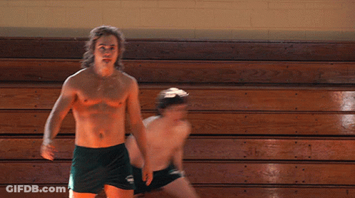 A Shirtless Man In Green Shorts Stands Next To Another Shirtless Man In Black Shorts GIF