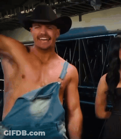 A Shirtless Man In Overalls And A Cowboy Hat Smiles GIF
