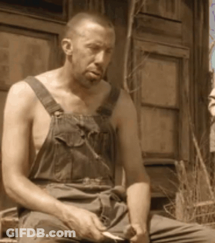 A Shirtless Man In Overalls Is Sitting In Front Of A Window GIF