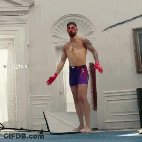 A Shirtless Man In Purple Shorts And Red Gloves Stands On A Blue Mat In A Room GIF