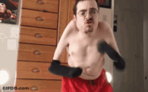 A Shirtless Man In Red Shorts And Black Gloves Is Standing In Front Of A Dresser . GIF