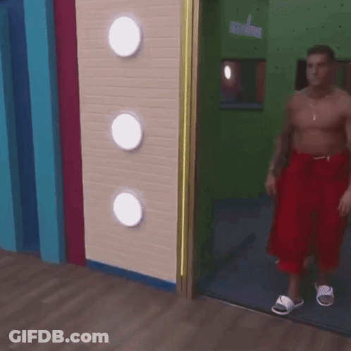 A Shirtless Man In Red Shorts And White Sandals Is Walking Through A Door GIF