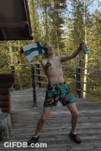 A Shirtless Man In Swim Trunks Is Holding A Flag And A Can Of Beer GIF