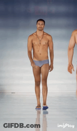 A Shirtless Man In Swim Trunks Walks Down A Runway At A Fashion Show GIF