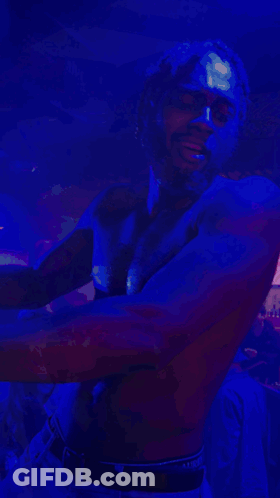 A Shirtless Man Is Dancing In A Dark Room With A Blue Light Behind Him GIF
