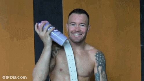 A Shirtless Man Is Holding A Purple Cup And Smiling While Wearing A White Belt . GIF