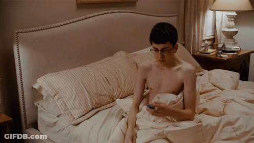 A Shirtless Man Is Laying On A Bed Looking At His Cell Phone GIF