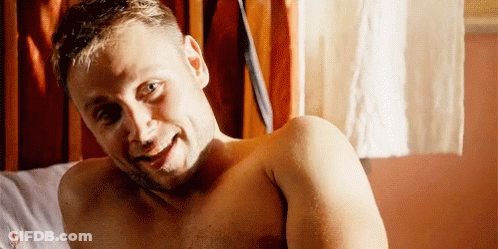 A Shirtless Man Is Laying On A Bed Smiling At The Camera . GIF