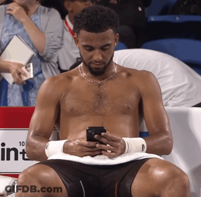 A Shirtless Man Is Looking At His Cell Phone In Front Of A Sign That Says 10 GIF