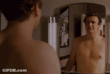 A Shirtless Man Is Looking At His Reflection In A Bathroom Mirror . GIF