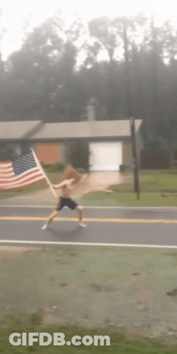 A Shirtless Man Is Running Down A Street With An American Flag In The Background GIF