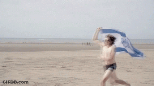 A Shirtless Man Is Running On The Beach Holding A Flag With A Blue And White Star On It GIF