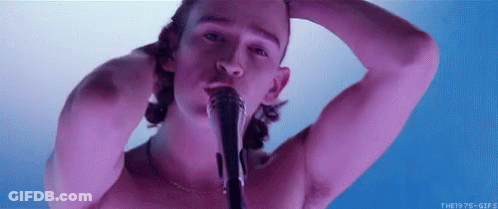 A Shirtless Man Is Singing Into A Microphone While Holding His Head . GIF