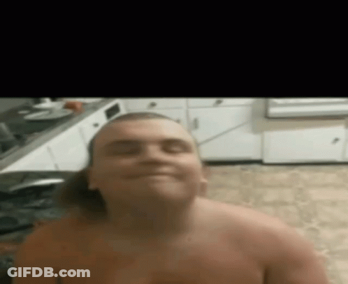 A Shirtless Man Is Sitting In A Kitchen And Smiling At The Camera . GIF
