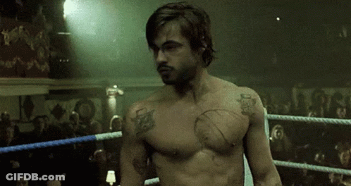 A Shirtless Man Is Standing In A Boxing Ring With A Crowd Watching . GIF
