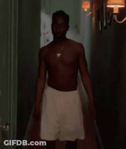 A Shirtless Man Is Standing In A Hallway Wearing White Shorts And A Necklace . GIF