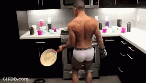 A Shirtless Man Is Standing In A Kitchen Holding A Pan . GIF