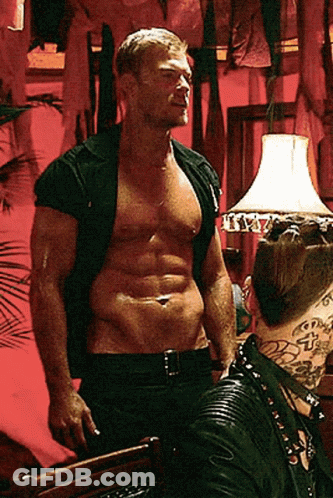 A Shirtless Man Is Standing In Front Of A Lamp With The Letter T On It GIF
