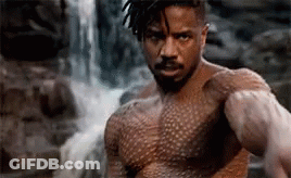 A Shirtless Man Is Standing In Front Of A Waterfall And Looking At The Camera . GIF
