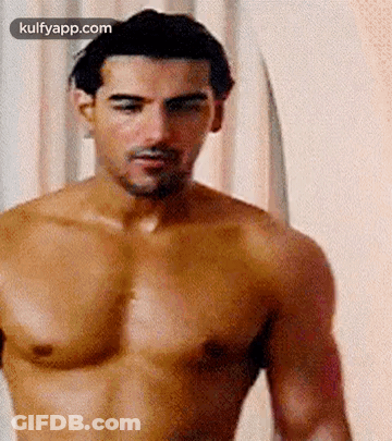 A Shirtless Man Is Standing In Front Of A White Curtain And Looking At The Camera . GIF