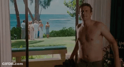A Shirtless Man Is Standing In Front Of A Window Looking Out At The Ocean . GIF