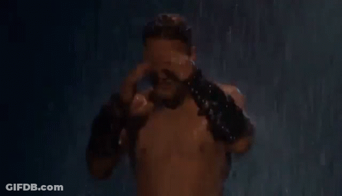 A Shirtless Man Is Standing In The Rain Holding His Head And Wearing Leather Gloves . GIF
