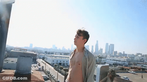 A Shirtless Man Is Standing On A Rooftop Overlooking A City Skyline . GIF