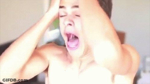 A Shirtless Man Is Yawning With His Mouth Open While Holding His Head In Front Of A Hair Dryer . GIF