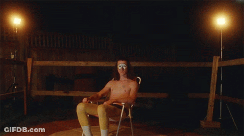 A Shirtless Man Sits In A Chair In Front Of A Wooden Fence At Night GIF