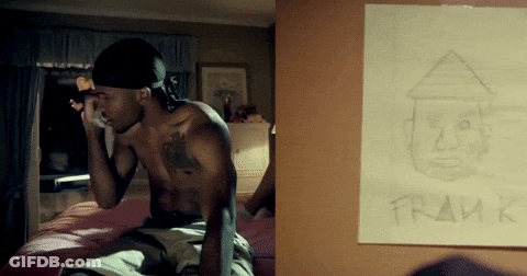A Shirtless Man Sits On A Bed Next To A Framed Drawing Of Him GIF