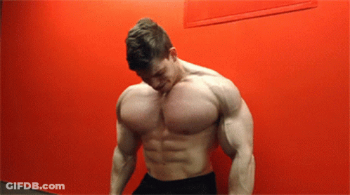 A Shirtless Man Stands In Front Of A Red Wall With His Head Down GIF