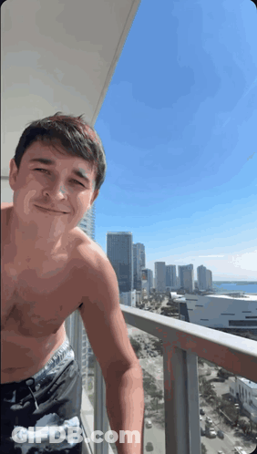 A Shirtless Man Stands On A Balcony With A City In The Background GIF