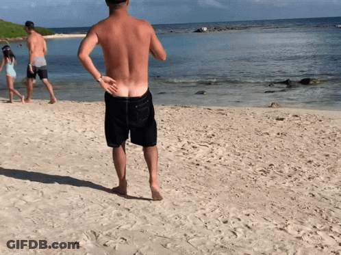 A Shirtless Man Stands On A Sandy Beach Looking At The Ocean GIF