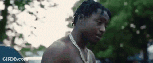 A Shirtless Man Wearing A Chain Around His Neck Is Standing In Front Of Trees . GIF