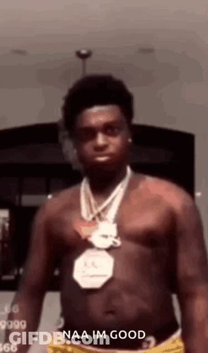 A Shirtless Man Wearing A Necklace And Shorts Is Standing In A Room With His Shirt Off . GIF