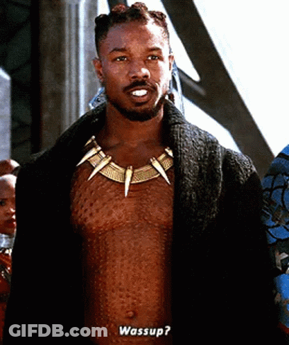 A Shirtless Man Wearing A Necklace With Sharp Teeth Is Standing In Front Of A Group Of People And Talking . GIF