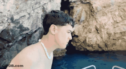 A Shirtless Man Wearing A Pearl Necklace Is Standing In Front Of A Body Of Water GIF
