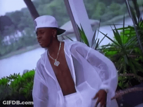 A Shirtless Man Wearing A White Hat And A Necklace With The Letter R On It Is Standing In Front Of A Body Of Water GIF